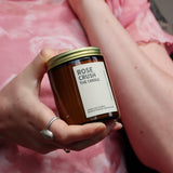 A person holding a amber glass jar candle with a gold brass lid and a label that reads 'ROSE CRUSH THE CANDLE'.