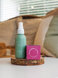 A hand holding a 50ml Fragrant Room Spray bottle with a green label, in front of a background of green leaves.