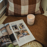 A small ginger-scented coconut wax candle with a lit flame, placed on an open magazine next to a brown-patterned pillow.