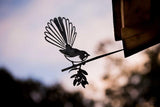 A metal wind spinner in the shape of a fantail or piwakawaka bird, with a fanned tail, mounted on a wall or roof at dusk.